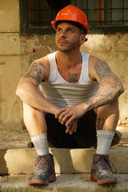 Man wearing hemp socks, shoes, shirt and shorts, as well as a hard hat sitting on steps with tattoos visible